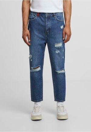 Vaqueros de mezclilla azul claro con detalles desgastados y rotos. Corte recto, con cierre de botones y trabillas para cinturón, combinados con zapatillas blancas.