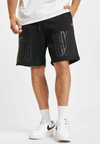 Fekete vászon sort, laza szabással, melyen a bal lábon egy nagy fekete "WEAR" felirat látható, valamint oldalsó zsebek találhatók. Fehér Nike tornacipővel viselik.