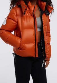 Pomarańczowa pikowana kurtka z wysokim kołnierzem, zapięciem na zamek i bocznymi kieszeniami. Posiada teksturowaną naszywkę na ramieniu oraz regulowane wykończenia na dole.