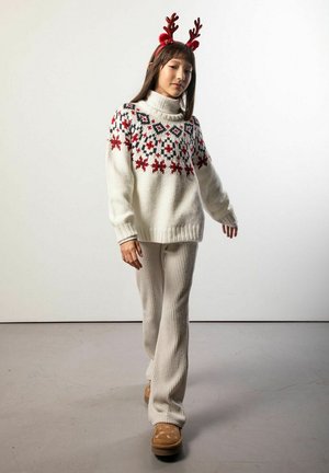 Biały dzianinowy sweter z czerwono-czarnymi geometrycznymi wzorami, zestawiony z kremowymi prążkowanymi spodniami i brązowym obuwiem. Modelka ma na głowie poroże renifera.