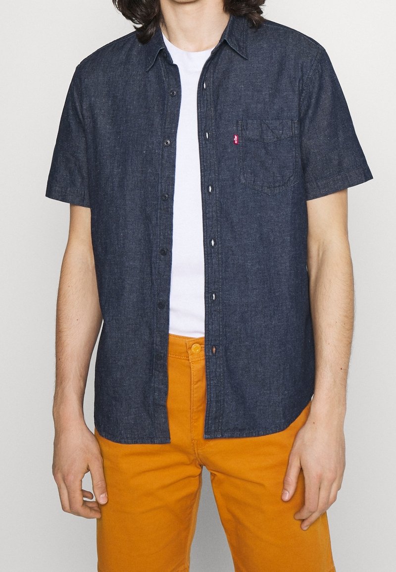 Kortärmad denimskjorta, mörkblå med knäppning och krage, med en bröstficka och Levi's-logotyp. Bärs med orange byxor.