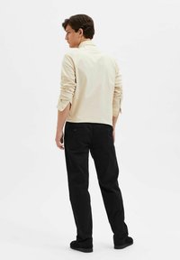 Beige long-sleeve shirt with rolled cuffs, paired with black pants. The outfit features a neutral color scheme and a relaxed fit.