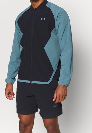 Homme portant une veste zippée Under Armour noire et bleu sarcelle et un short noir Nike, debout devant un fond uni.