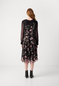 Schwarzes Blumenmusterkleid mit langen transparenten Ärmeln, einem kragenförmigen Ausschnitt und einem fließenden Saum, der mit rosa und grünen Blumenmustern dekoriert ist. Schwarze Knöchelschuhe.
