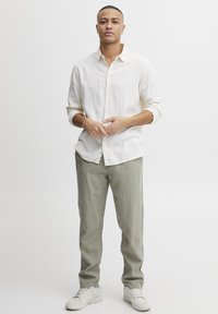 Camicia button-up di lino beige chiaro, maniche arrotolate, abbinata a pantaloni verde oliva chiaro e sneakers bianche. Il modello si trova in piedi con le mani intrecciate.