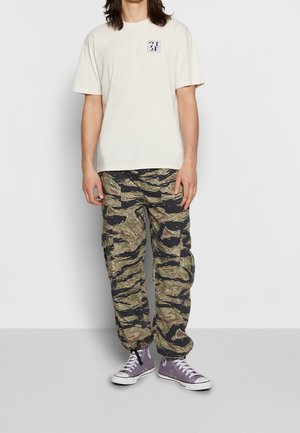 Camiseta de algodón beige con un pequeño logo azul en el pecho, combinada con pantalones cargo de camuflaje con patrones verdes y negros. Zapatillas moradas.