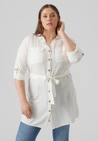 Camisa blanca de botonadura con dos bolsillos en el pecho y mangas enrolladas, con un cinturón anudado en la cintura y botones dorados. Textura de tela suave.