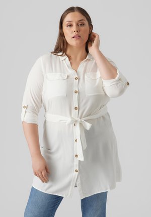 Camisa blanca de botonadura con dos bolsillos en el pecho y mangas enrolladas, con un cinturón anudado en la cintura y botones dorados. Textura de tela suave.