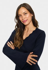 Donna con lunghi capelli castani mossi che indossa un maglione blu navy a scollo a V con maniche svasate, braccia incrociate, sorridente delicatamente su uno sfondo bianco.