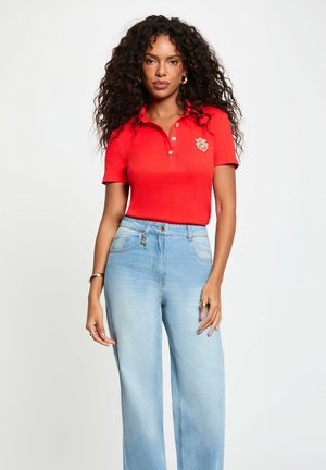 Donna con capelli ricci che indossa una polo rossa a maniche corte con stemma e jeans a vita alta e gamba larga azzurri chiari su sfondo bianco.