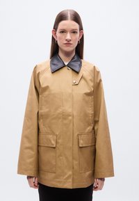 Καφέ αδιάβροχο μπουφάν με μαύρο γιακά, μπροστινό κούμπωμα με τρουκ και δύο τσέπες επιδιόρθωσης. Διαθέτει άνετα μανίκια και λεία υφή.