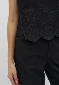 Zwarte cropped top met geborduurde ronde patronen, een scalloped zoom, gecombineerd met zwarte gestructureerde high-waisted broeken.