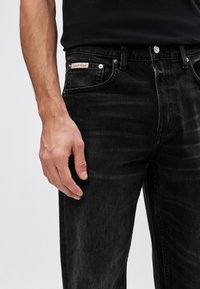 Jeans din denim negri cu o textură estompată, buzunar frontal, închidere cu nasture și o etichetă cu logo aproape de talie. O mână se odihnește lângă buzunar.