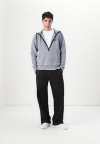 Sudadera gris con cremallera y patrón a cuadros, detalles negros y bolsillos frontales, llevada sobre una camiseta blanca y combinada con pantalones negros y zapatos blancos.