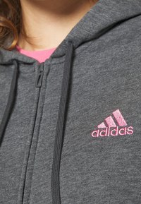 Sweat à capuche gris avec logo Adidas rose. Doté d'une fermeture éclair et de cordons de serrage. Texture lisse, design décontracté et couche intérieure rose contrastante.