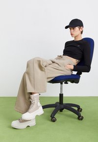 Pantalons de survêtement beiges surdimensionnés, top noir à manches longues et bottes à chevilles chunky de couleur crème avec des semelles texturées. Assis sur une chaise de bureau bleue.