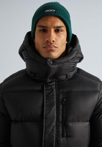 Schwarze Pufferjacke mit hohem Kragen, glänzender Textur und einer seitlichen Reißverschluss-Tasche. Getragen mit einer grünen Strickmütze mit Logo.