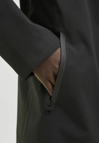 Detaliu al unui hanorac impermeabil negru, cu mâna în buzunarul cu fermoar. Textură netedă, croi ajustat și design cu fermoar ascuns.