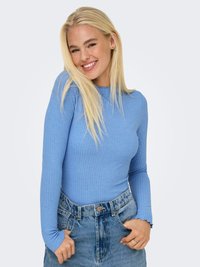 Jauna sieviete ar gariem blondiem matiem, kura ir ģērbusies gaiši zilā garroku krekliņā un augsta vidukļa džinsos, smaida ar rokām kabatās.