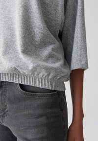 Gros plan sur une personne portant un pull en tricot gris clair avec un ourlet élastique et un jean gris foncé, montrant une partie du torse et du bras droit.