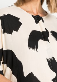 Czarny i beżowy top z abstrakcyjnymi wzorami przypominającymi pociągnięcia pędzla, wykonany z miękkiego materiału, z zaokrąglonym dekoltem i luźnymi krótkimi rękawami. Minimalistyczny design.
