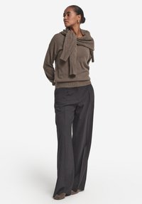 Brauner Strickpullover mit drapiertem Kragen, kombiniert mit dunkler, maßgeschneiderter Hose. Das Outfit zeichnet sich durch eine lockere Passform und weiche Textur aus, dazu passende Schuhe.