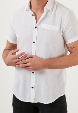 Uomo con una camicia bianca a maniche corte con texture e bottoni neri e tatuaggi su entrambi gli avambracci, mani incrociate davanti.