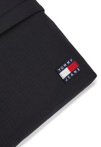 Μαύρη υφασμάτινη θήκη σε ορθογώνιο σχήμα με υφή επιφάνειας. Διαθέτει ραμμένη ετικέτα "TOMMY JEANS" με κόκκινα, λευκά και μπλε χρώματα.