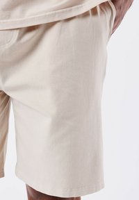 Pantaloni corti in cotone beige con una texture liscia, dotati di tasche laterali e una vestibilità rilassata. L'orlo è dritto e termina appena sopra il ginocchio.