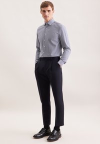 Uomo in piedi con le mani nelle tasche, indossa una camicia a maniche lunghe a quadretti, pantaloni eleganti scuri, calzini neri e scarpe da uomo in pelle nere.