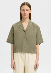 Olivově zelená, zkrácená košile na knoflíky s klopovým límcem, krátkými rukávy a kapsou na levé straně hrudi. Hladká tkanina a minimální detaily.
