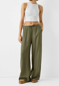 Bílý žebrovaný crop top a volné olivově zelené kalhoty se strečovým pasem a detailem na zavazování. Viditelné hnědé platformové sandály.