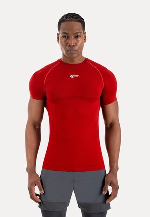 Muskuløs mand med flettet hår iført stram rød sportstrøje og grå shorts, stående med fronten mod kameraet foran en hvid baggrund.