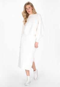 Pull en tricot blanc avec un grand col et des manches ballon, associé à une jupe longue et droite assortie avec une fente latérale et des baskets blanches.