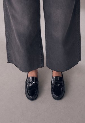 Persoon die een brede donkere spijkerbroek en glanzende zwarte leren loafers draagt, staand op een grijze vloer.