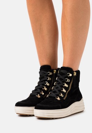 Sorte ruskindssko med plateau og guldknapper, side lynlåse, sorte snørebånd, båret på bare ben mod en hvid baggrund.