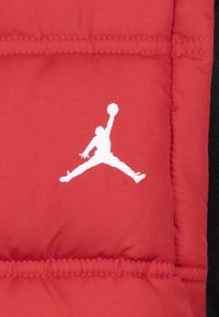 Rød quiltet stof med syet detaljer har et hvidt Jumpman-logo, der tydeligt vises. Teksturen er glat og fyldig.