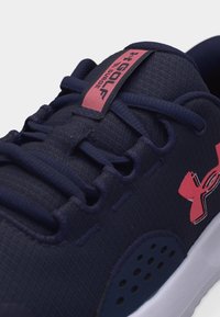 Κοντινό πλάνο μπλε παπουτσιού γκολφ Under Armour με ροζ λογότυπο και ετικέτα "Golf Surge" στη γλώσσα, που δείχνει υφασμάτινη υφή και κορδόνια.