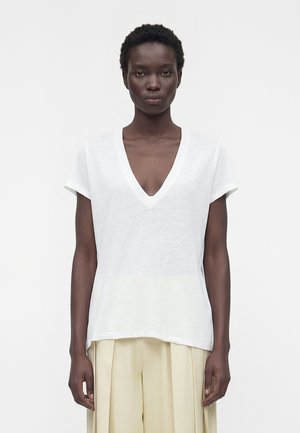 Femme aux cheveux courts et bouclés portant un haut blanc à manches courtes en V et un pantalon beige plissé à jambes larges, debout devant un fond uni.