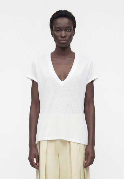 Frau mit kurzen lockigen Haaren, die ein weißes V-Ausschnitt-Kurzarmshirt und beige plissierte weite Hosen trägt, vor einem schlichten Hintergrund stehend.
