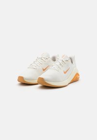 Balti sportiniai bateliai su oranžiniais akcentais, tekstūruota tinklinė viršutinė dalis, guminis padas ir ryškus Nike logotipas šone ir liežuvyje.