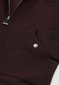 Hanorac cu fermoar de culoare burgundy, din material neted; are o buzunar mare cu un accent de nasture argintiu și detalii de cusătură subtile.