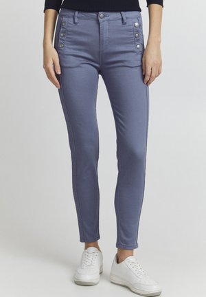 Slim fit, hooggewaisted blauwe jeans met een gladde textuur. Voorzien van grote zilveren knopen aan de zijkanten en contrasterende stiksels. Gepaard met witte sneakers.