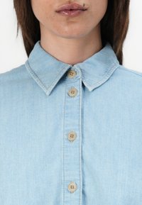 Chemise en jean bleu clair avec un col pointu, patte de boutonnage avant et six boutons beiges, présentant une texture lisse et un design décontracté.