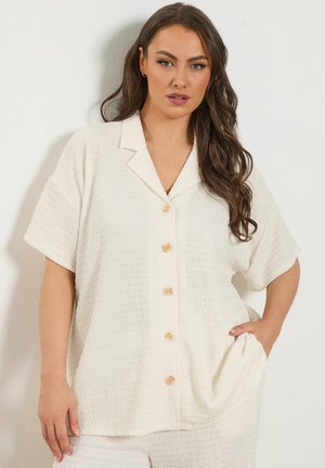 Frau mit langem braunem Haar trägt eine strukturierte, weiße Kurzarm-Hemdbluse mit Kragen und passenden Hosen und posiert mit einer Hand in der Tasche.