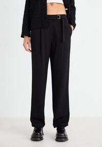 Pantalones negros ajustados con un diseño de pierna recta, que incluyen un cinturón con una hebilla plateada y bolsillos laterales. Llevados con calzado negro.