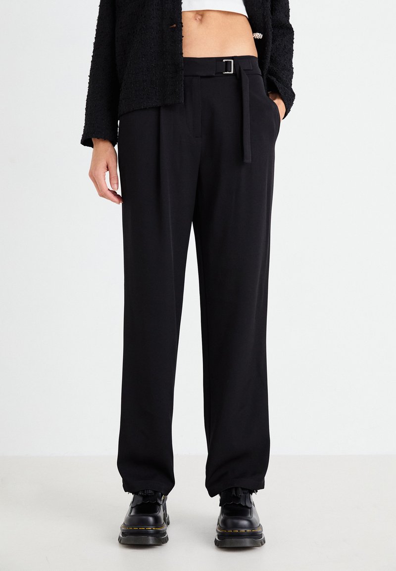 Pantalones negros ajustados con un diseño de pierna recta, que incluyen un cinturón con una hebilla plateada y bolsillos laterales. Llevados con calzado negro.