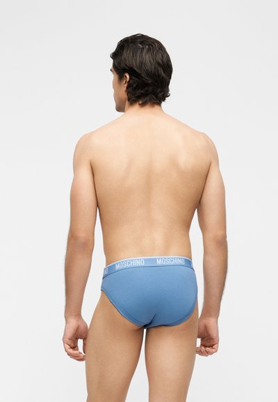 Άνδρας με σκούρα μαλλιά στέκεται με την πλάτη, φορώντας γαλάζια εσώρουχα Moschino μπροστά σε απλό γκριανο υπόβαθρο.