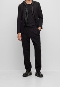 Outfit für schwarze Männer mit einem maßgeschneiderten Blazer mit strukturierter Kapuzenfutter, einem gestrickten schwarzen Shirt und gemusterten schwarzen Hosen. Schwarze Schuhe runden das Outfit ab.