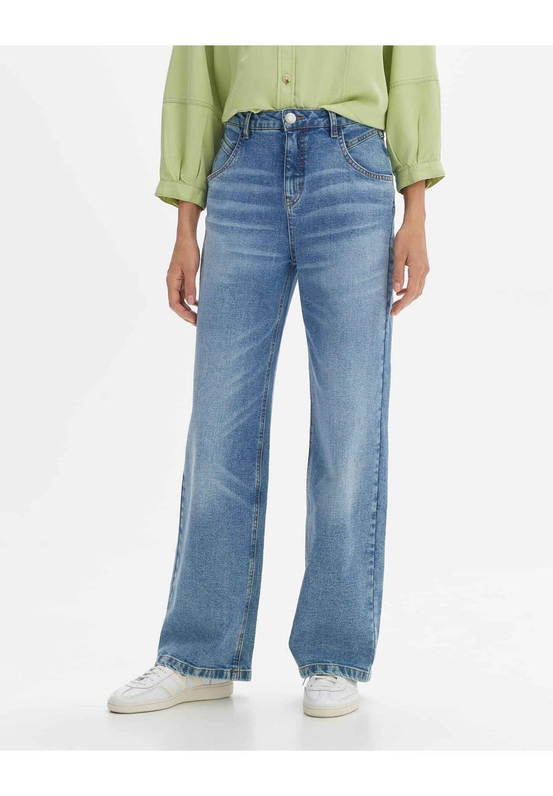 Light blue denim wide-leg jeans feature a high waist, five pockets, and subtle fading. Paired with white sneakers, and a green top.